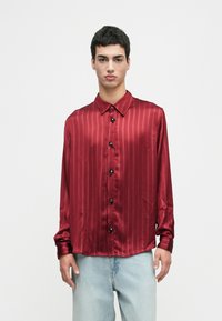 Young man wearing a red satin striped long-sleeve shirt with decorative black buttons paired with light blue jeans against white background.