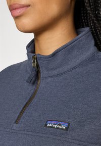 Detailný záber na osobu, ktorá má na sebe modrý fleecový sveter Patagonia s polovičným zipsom a viditeľným logom Patagonia na hrudi.