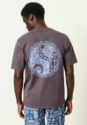 Mand iført mørkegrå t-shirt med cirkulært pool-tema grafik og tekst på ryggen, kombineret med blå mønstrede shorts.