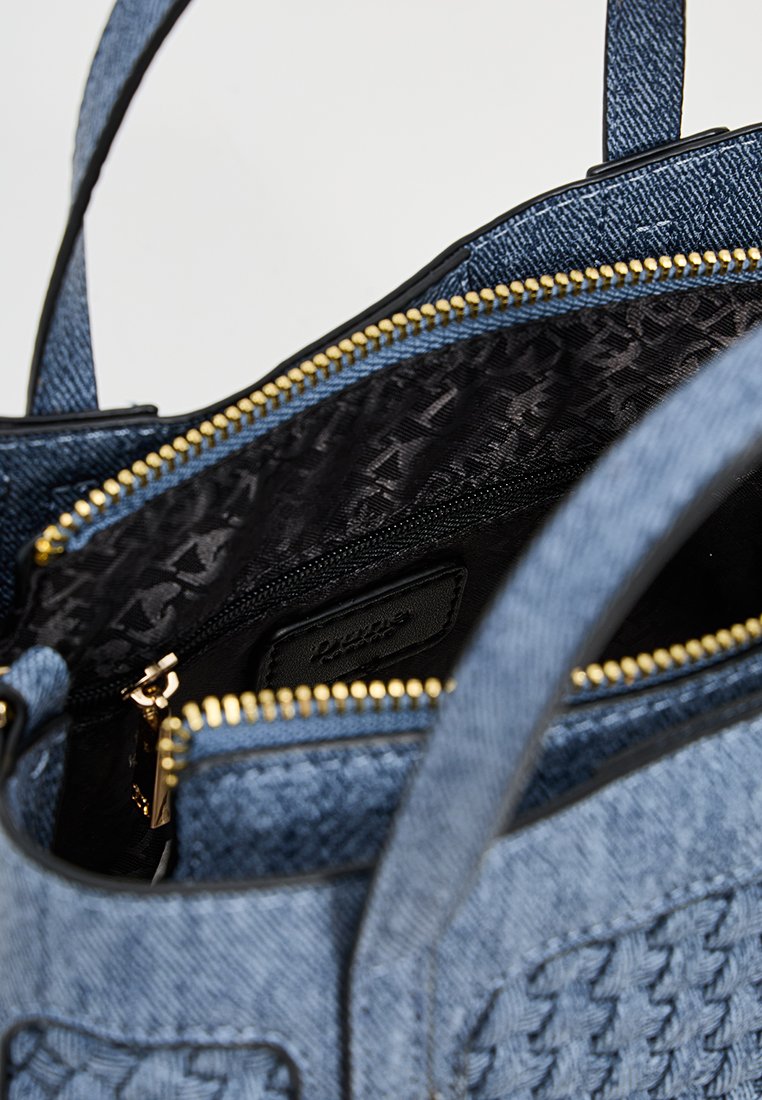Sac fourre-tout en denim avec une fermeture éclair dorée, présentant un intérieur noir à motif et une poche zippée. L'extérieur est doté de détails tissés.