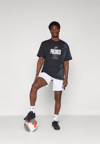 Nike Premier sort grafisk t-shirt med hvid tekst, parret med hvide shorts. Modellen står på en fodbold, iført sorte støvler og hvide strømper.
