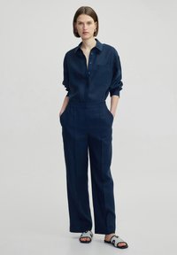 Marineblauer Jumpsuit mit einem Kragen, langen Ärmeln, seitlichen Taschen und einer lässigen Passform. Verfügt über Knopfdetails auf der Brust.
