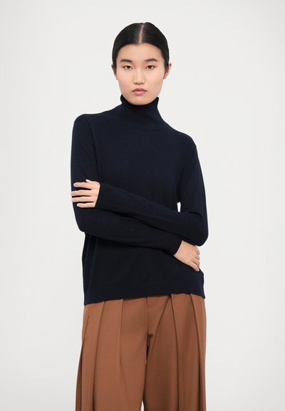 Navy blue turtleneck sweater made from soft fabric, featuring a relaxed fit and long sleeves, paired with brown pleated trousers.