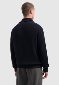 Man die met zijn rug naar de camera staat, draagt een donkerblauwe geribde trui met lange mouwen en grijze broek tegen een effen lichte achtergrond.