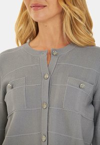 Cardigan grigio chiaro a trama con scollatura rotonda, dotato di decorativi bottoni e due tasche sul petto.