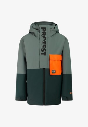 Garçon debout portant une veste verte et grise avec une poche orange, un pantalon orange, des gants noirs, des bottes noires et un bonnet en laine noir.