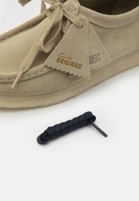 Mocassino in suede beige con chiusura a lacci, caratterizzato da un'etichetta in pelle con la scritta "Clarks Originals" e un accessorio in pizzo color navy accanto ad essa.