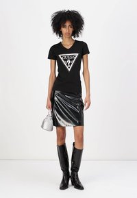T-shirt nera con logo a triangolo e dettagli in strass. Gonna in vinile nero lucido, borsa argentata e stivali neri al ginocchio.