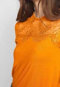 Oranžový top s krátkým rukávem a krajkovým panelem u výstřihu. Hladká textura, těsný střih a jemný lesk na látce.