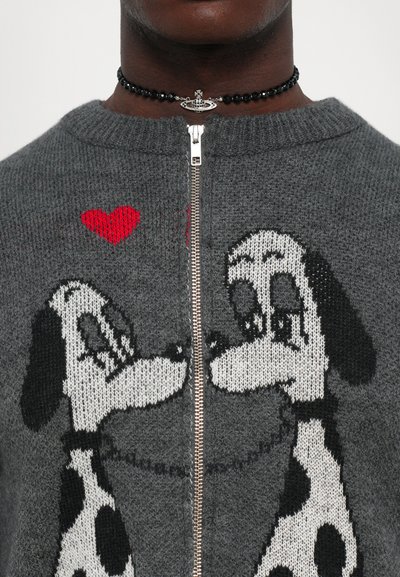 Detailaufnahme eines grauen Reißverschluss-Pullovers mit zwei schwarz-weißen Cartoon-Hunden, die sich gegenüberstehen, und einem roten Herz über ihnen, getragen von einer Person mit einer Perlen-Halskette.
