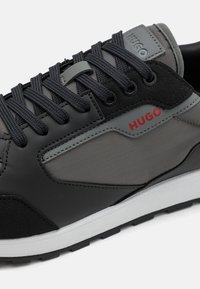 Zwart en grijze sneaker met een glad bovenwerk van synthetisch materiaal, textuuraccenten, contrasterend logo en een witte zool met een robuust profiel.