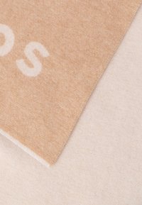 Tan uldblandet stof med blød tekstur, med hvid tekst på en sektion, pænt syede kanter og en beige baggrund.