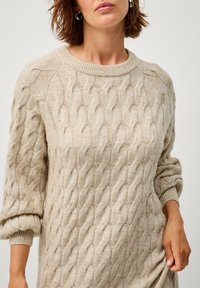 Beige Kabelstrickpullover mit langen Ärmeln, geripptem Halsausschnitt und Bündchen, ausgestattet mit einem strukturierten Muster und weichem, warmem Stoff.