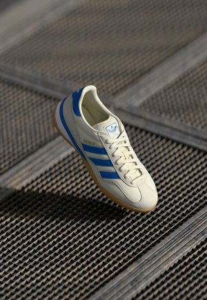 Adidași albi cu dungi și accente albastre, design cu șireturi și talpă din cauciuc natural. Dispune de un material superior texturat și logo pe limba pantofului.