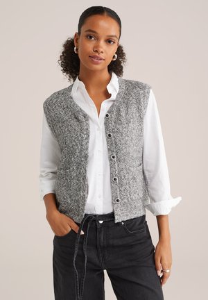 Gilet tricoté gris texturé avec un col en V et une fermeture à boutons sur une chemise blanche à manches longues. Un jean noir complète la tenue.
