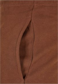 Bruine rekbare stof met een gladde textuur, voorzien van een zijzak geïntegreerd in het ontwerp, gestikt met subtiele naden.