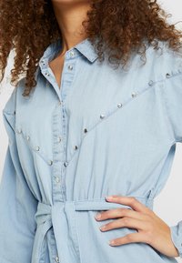 Light blue denim shirt with a slim fit, featuring snap buttons along the front and diagonal accents, styled with a tied belt at the waist.