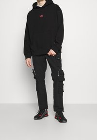 Sweat à capuche noir avec une poche avant et une petite étiquette rouge. Pantalon cargo noir avec plusieurs poches et fermetures éclair. Chaussures noires avec des accents rouges.