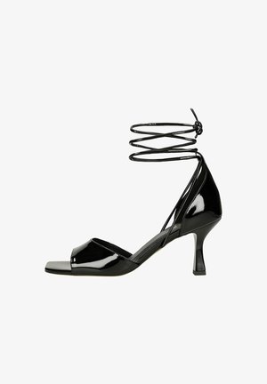 Czarne lakierowane sandały z otwartym palcem, zawiązywane wokół kostki i cienkim, zakrzywionym obcasem. Zawierają płaski przód i teksturowane paski.