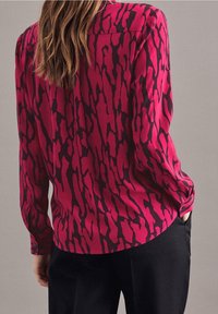 Donna con lunghi capelli castani che indossa una blusa magenta a maniche lunghe con motivi astratti neri e pantaloni neri, con una mano in tasca.
