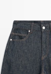 Dunkelblaue Jeans mit Fronttaschen, Gürtelschlaufen, Metallnieten und Knopfverschluss, von der Taille bis zur Mitte des Oberschenkels auf weißem Hintergrund gezeigt.