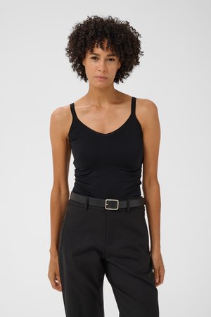 Femme aux cheveux bouclés portant un top noir sans manches, un pantalon noir et une ceinture noire, debout devant un fond clair uni.