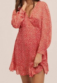 Rotes Blumenkleid mit langen Ärmeln, V-Ausschnitt und Rüsche am Saum. Der Stoff weist ein weißes Blumenmuster auf einem strukturierten Hintergrund auf.