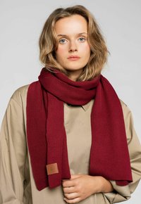 Bordowy, dziergany szalik wykonany z teksturowego materiału, z skórzaną metką, zarzucony na szyję modelki noszącej beżową bluzkę.