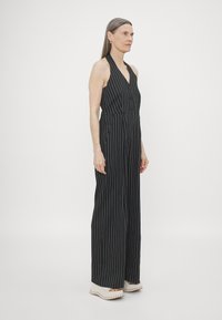 Svartstripet jumpsuit med halterneck, frontknapper og vide ben. Stoffet ser glatt ut, og designet har vertikale hvite striper.