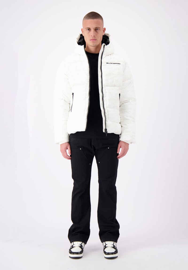 Witte pufferjack met een gestructureerde afwerking, zwarte ritsen en logo-detail. Gecombineerd met zwarte broek en sneakers met een patroon.