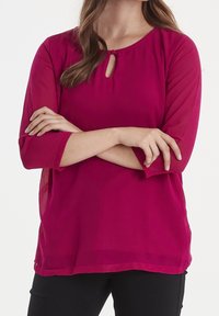 Blusa fucsia con maniche a tre quarti, scollo rotondo e dettaglio a chiave. Tessuto leggero con una texture liscia e una vestibilità comoda.