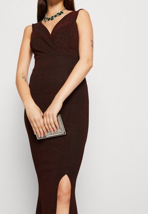 Vrouw draagt een mouwloze donker bordeauxrode jurk met een voorsplit, houdt een zilveren clutch vast en draagt een groene edelstenen ketting.