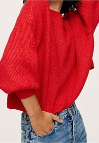 Un pull rouge en tricot avec une texture côtelée, des manches ballon et un col rond, associé à un jean en denim bleu taille haute.