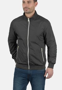 Schwarze Bomberjacke für Herren aus leichtem Material. Sie verfügt über einen Stehkragen, einen Reißverschluss an der Vorderseite, gerippte Bündchen und seitliche Taschen mit weißen Reißverschlüssen.
