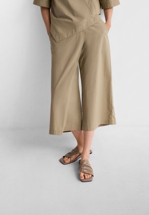 Pantaloni larghi color khaki realizzati in tessuto leggero, abbinati a sandali marroni con cinturini incrociati e suola piatta e quadrata.