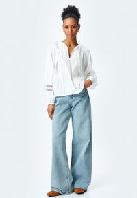 Hvid bomuldsbluse med intrikate blonder, løstsiddende pasform og lange ærmer, parret med blege blå bukser med bred ben og brune sko.