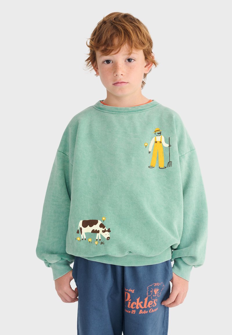 Garçon aux cheveux roux portant un sweat-shirt vert avec une illustration d'agriculteur et de vache, un pantalon bleu avec le texte orange "Pickles" et un motif de chien.