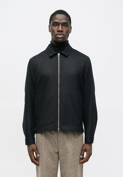 Man wearing a black zip-up jacket over a black turtleneck and beige textured pants, standing against a plain light background.