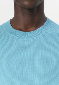 Svetlo modra pletena majica z teksturirano površino in okroglim izrezom. Tkanina se zdi mehka in zračna, primerna za vsakodnevno nošenje.