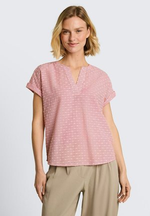 Femme aux cheveux blonds courts portant un chemisier rose texturé à manches courtes et un pantalon beige, debout avec un léger sourire sur un fond uni.