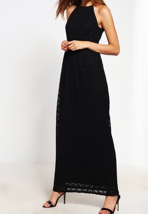 Vrouw draagt een mouwloze zwarte maxijurk van kant met halternek en zwarte sandalen met hoge hakken, staand tegen een effen witte achtergrond.