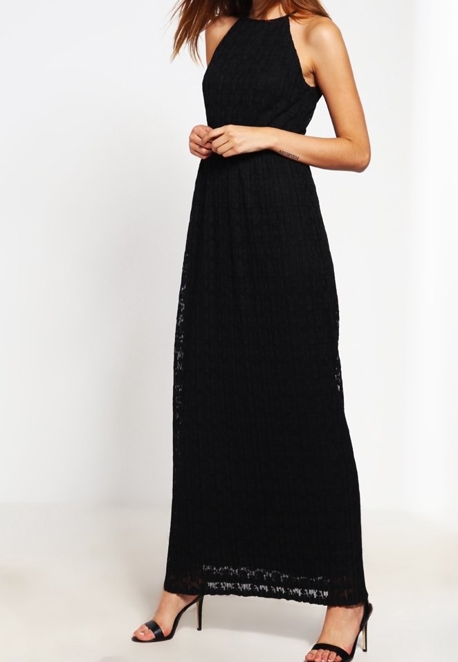 Femme portant une robe maxi sans manches en dentelle noire avec un col halter et des sandales noires à talons hauts, debout devant un fond blanc uni.