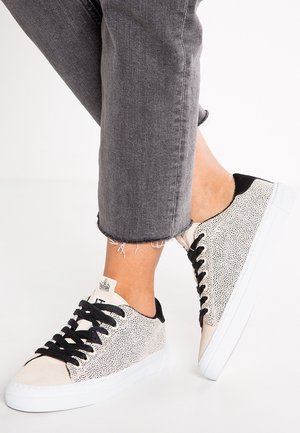 Person iført grå ankellange jeans og hvide lavtop sneakers med sorte pletter og sorte snørebånd på hvid baggrund.