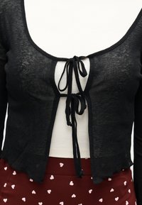 Μαύρο διαφανές καρντίγκαν με κορδόνια πάνω από λευκό ριμπ τοπ, συνδυασμένο με μπορντό φούστα με λευκά σχέδια καρδιών.