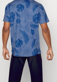 Sininen lyhythihaisia paita, jossa on trooppinen lehti- ja kukkakuosi. Valmistettu tekstuurista kankaasta, jossa on rento leikkaus ja sivuhalkiot.