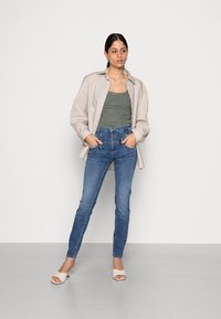 Camisa oversized bege, top de alças verde com padrão, jeans azul de cintura alta e sapatos de salto abertos brancos, posando num fundo cinza claro.