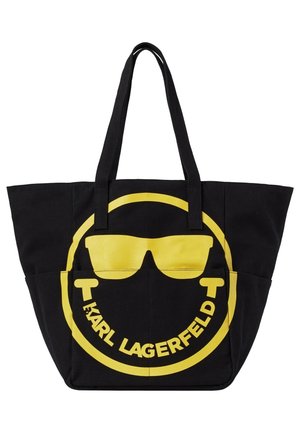 Sac cabas noir avec un visage souriant jaune portant des lunettes de soleil et le texte « Karl Lagerfeld » courbé sous le visage à l'intérieur d'un cercle jaune.