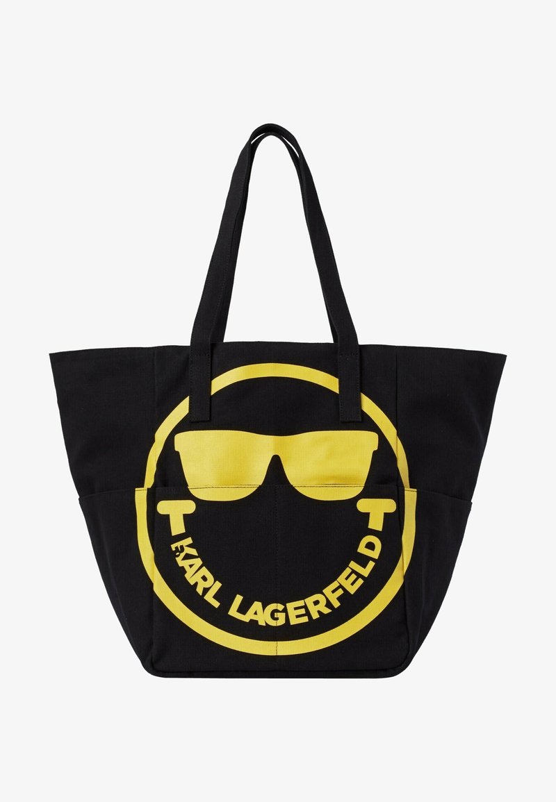 Sac cabas noir avec un visage souriant jaune portant des lunettes de soleil et le texte « Karl Lagerfeld » courbé sous le visage à l'intérieur d'un cercle jaune.