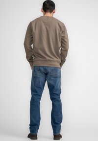 Bruine sweatshirt met ribgebreide manchetten en onderkant, gecombineerd met blauwe spijkerbroek. De outfit wordt compleet gemaakt met donkere schoenen.