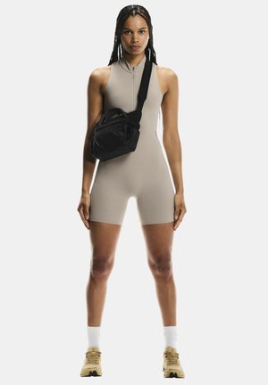 Woman with braided hair wearing beige sleeveless zip-up romper, black crossbody bag, white socks, and beige sneakers, standing facing forward.